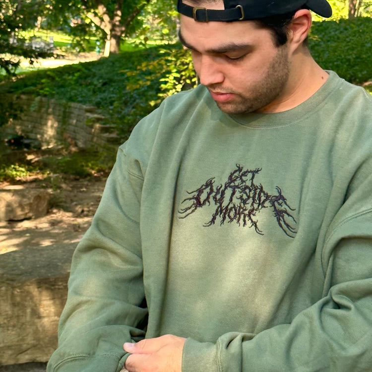 A man wearing an olive green sweatshirt with the words 'Get Outside More' embroidered on it in a metal band font. Shop "Get Outside More," an edgy nature clothing brand featuring outdoor-themed shirts.