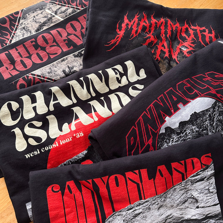 A pile of shirts depicting imagery of various national parks. Shop our National Park Graphic Tees, the ultimate national parks apparel! Edgy, nature-themed designs inspired by band merch, perfect for showcasing your love for adventure.