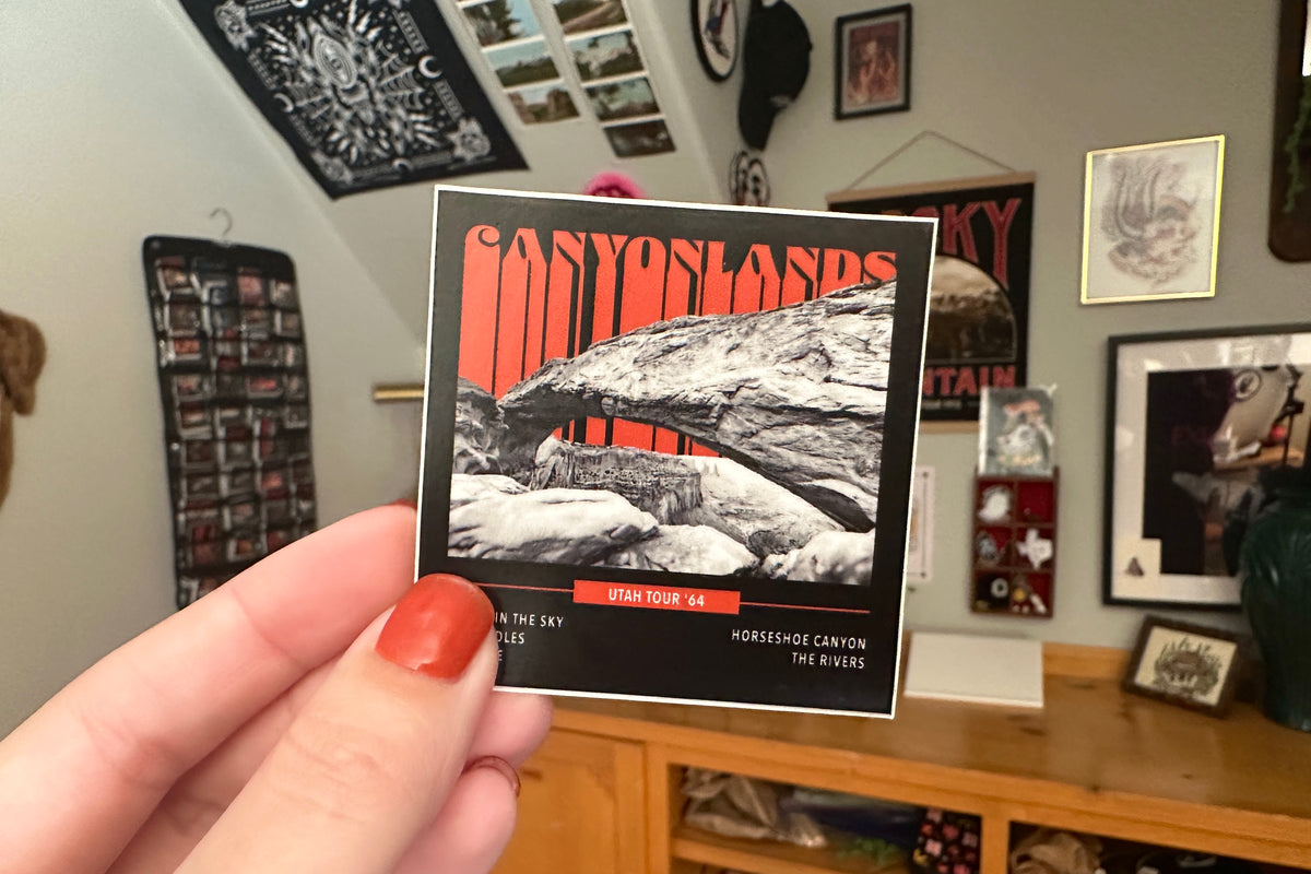 Person holding a 'Canyonlands' sticker in a room with framed pictures on the wall.