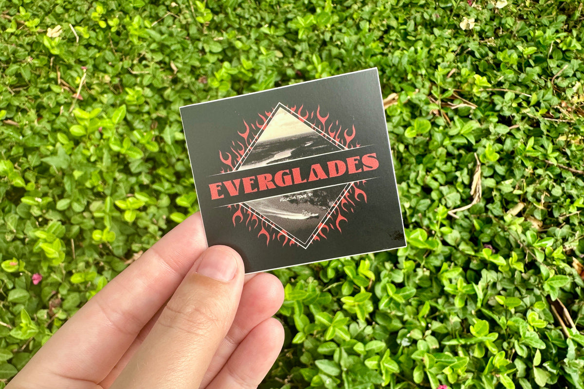 Hand holding a black 'Everglades' sticker against a green grass background
