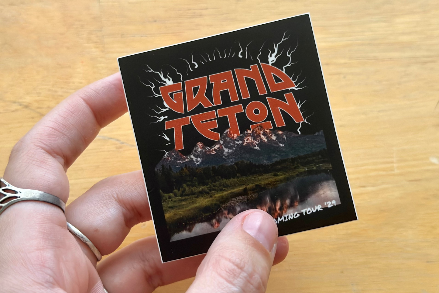Hand holding a sticker with 'Grand Teton' text and image on a wooden surface