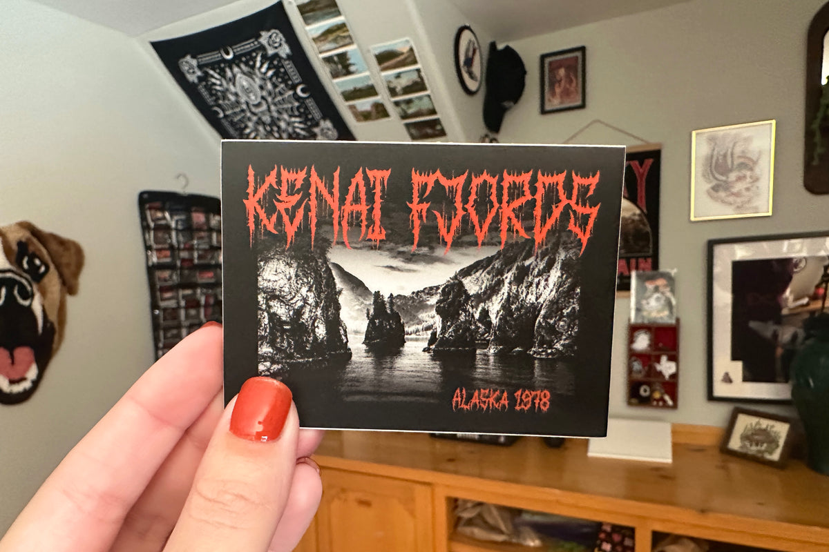 Person holding a 'Kenai Fjords' sticker in a room with framed pictures on the wall.
