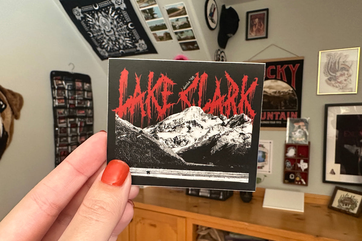 Hand holding a sticker with 'Lake Clark' design in a room with framed pictures on the wall.