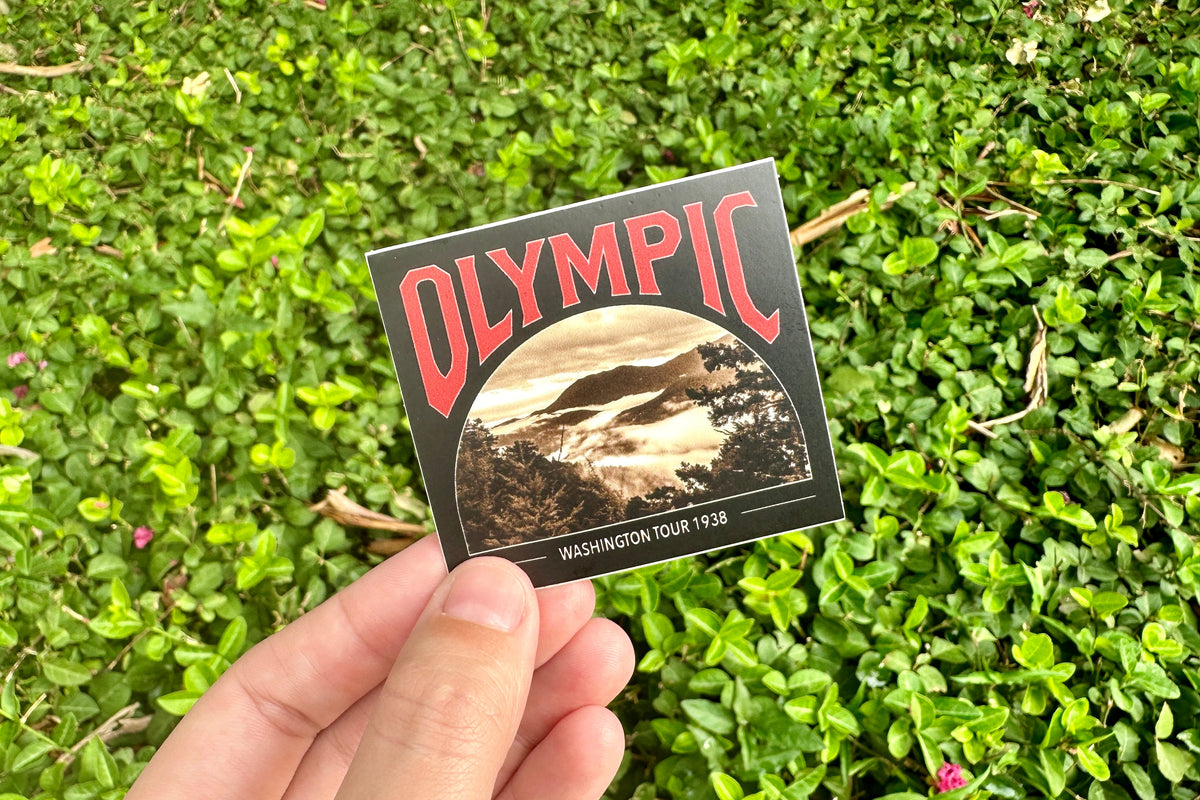 Hand holding a 'Olympic' vintage-style sticker against a grassy background