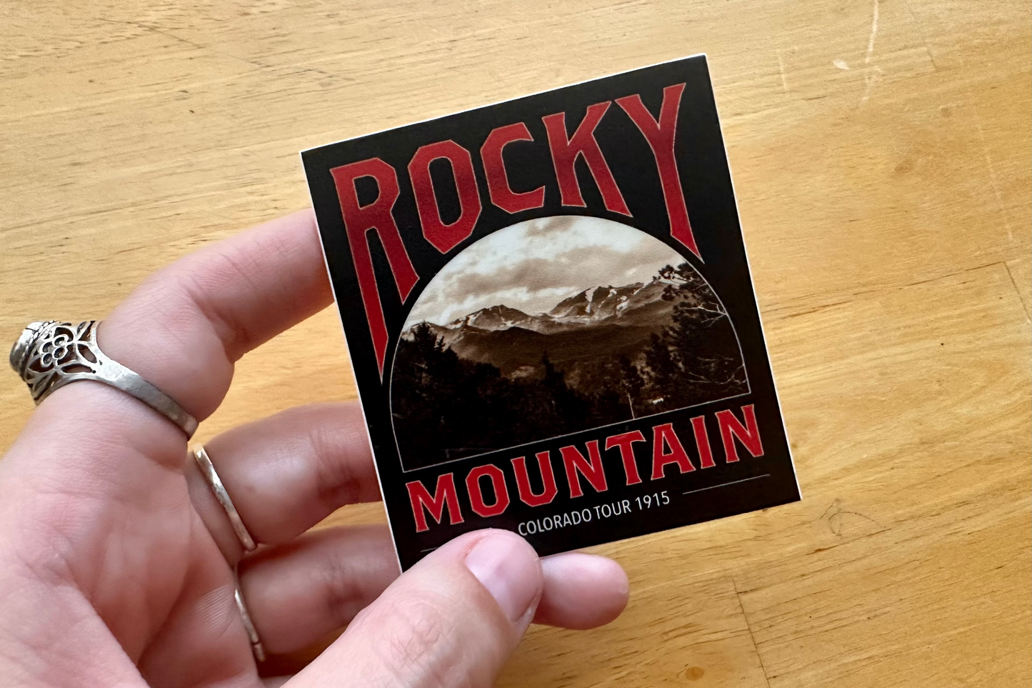 Hand holding a Rocky Mountain sticker on a wooden surface