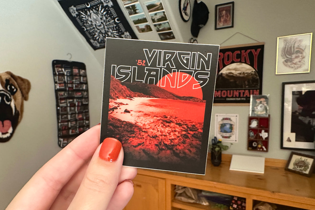 Person holding a 'Virgin Islands' sticker in a room with various posters and decor.
