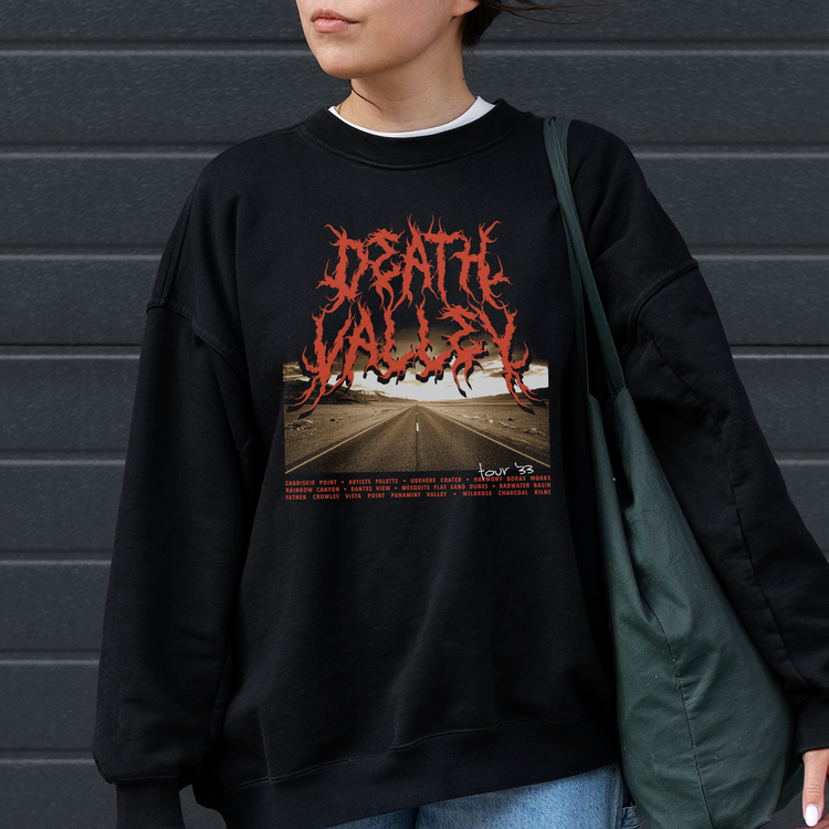 A woman wearing a black sweatshirt with the words 'Death Valley' on it in a metal band font. Shop our National Park Sweatshirts, the perfect national parks apparel! Featuring unique designs inspired by iconic parks, they combine comfort and adventure in s