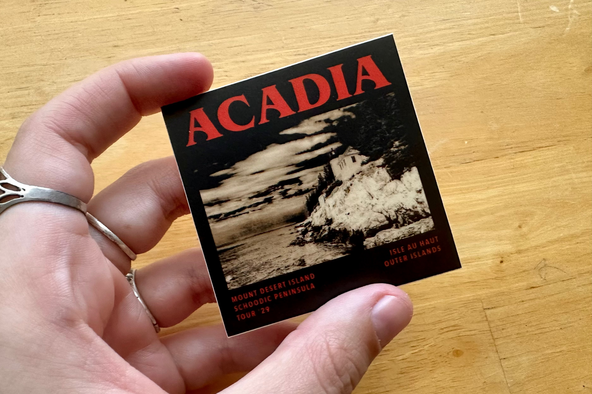 Hand holding a sticker with 'Acadia' design on a wooden surface