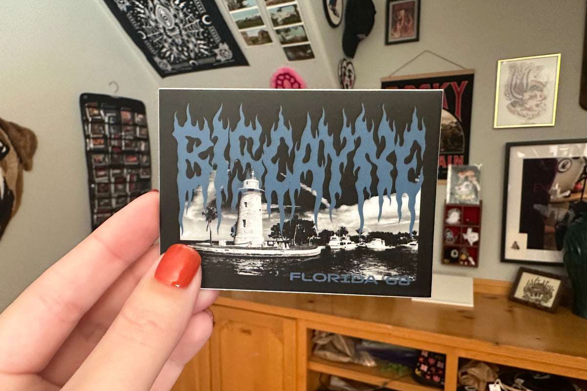 Person holding a 'Biscayne' sticker in a room with framed pictures on the wall.