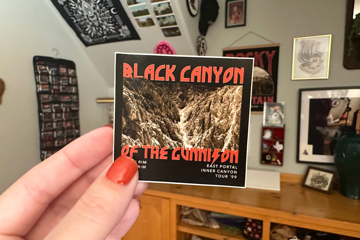 Hand holding a 'Black Canyon of the Gunnison' sticker in a room with framed pictures on the wall.
