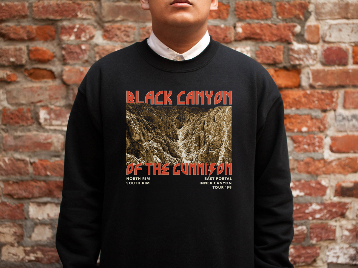 A person wearing a black crewneck sweatshirt with 'Black Canyon of the Gunnison' text and an image of a mountain landscape on the front, standing in front of a brick wall.