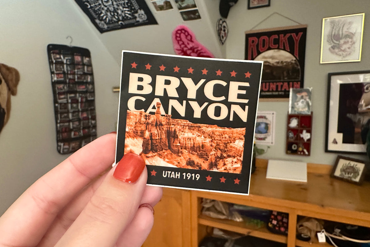 Hand holding a 'Bryce Canyon' sticker in a room with posters on the wall.