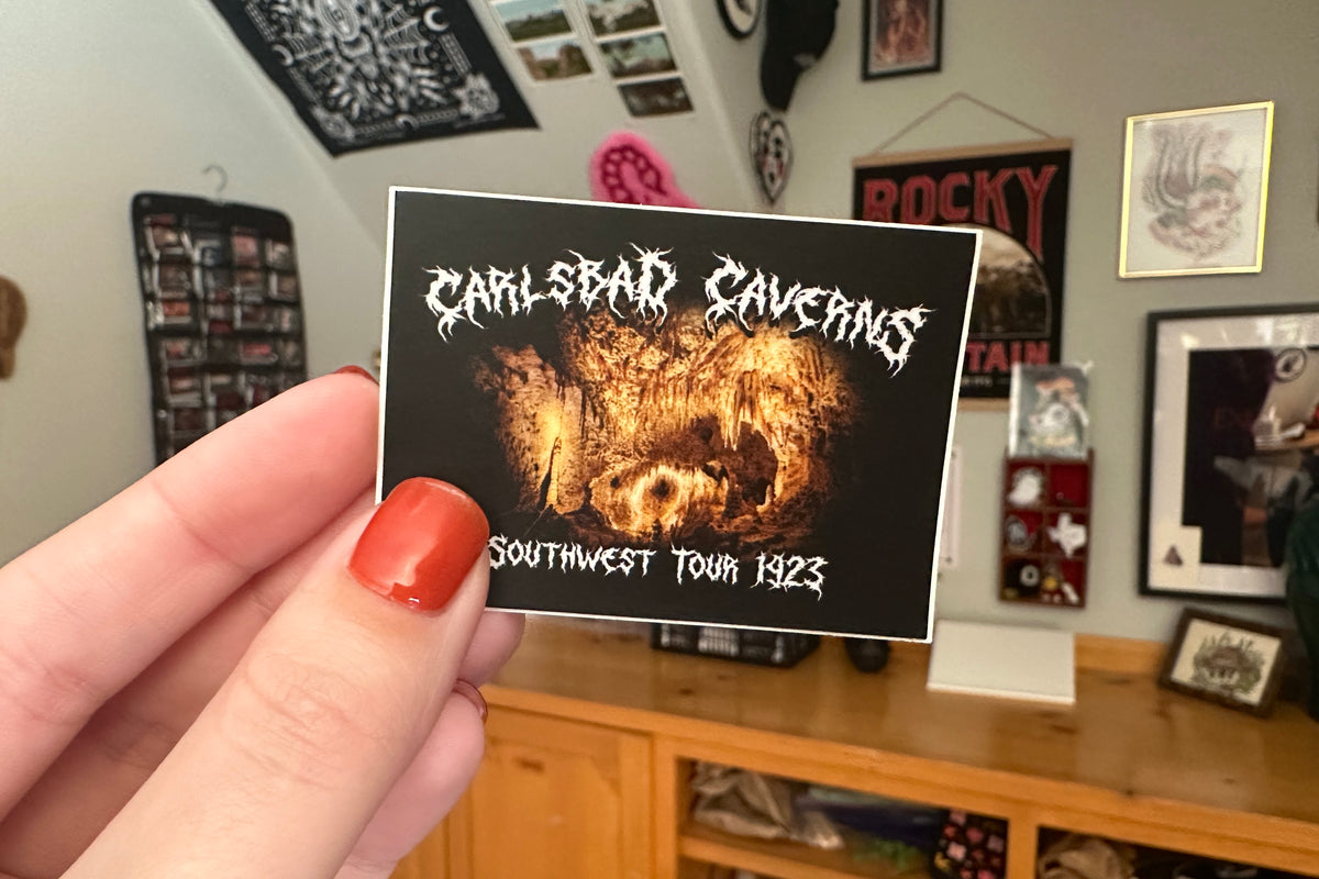 Hand holding a 'Carlsbad Caverns' sticker in a room with posters on the wall.