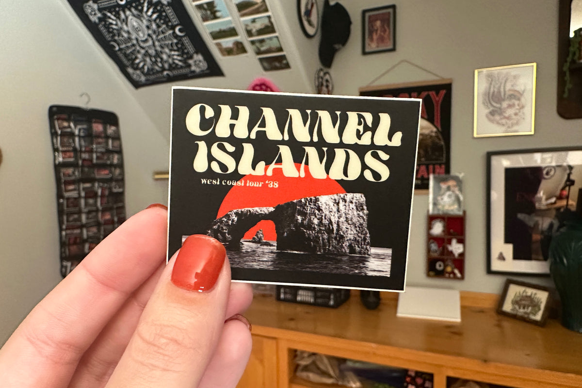 Hand holding a 'Channel Islands' sticker in a room with various framed pictures on the wall.