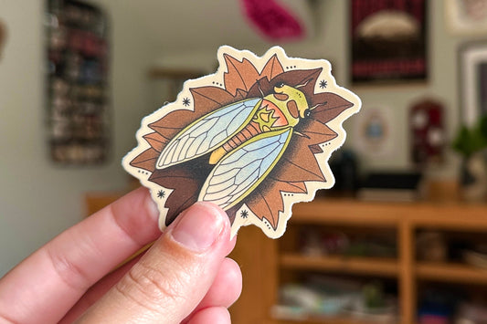 Hand holding a decorative sticker with a stylized insect design in front of a blurred indoor background.