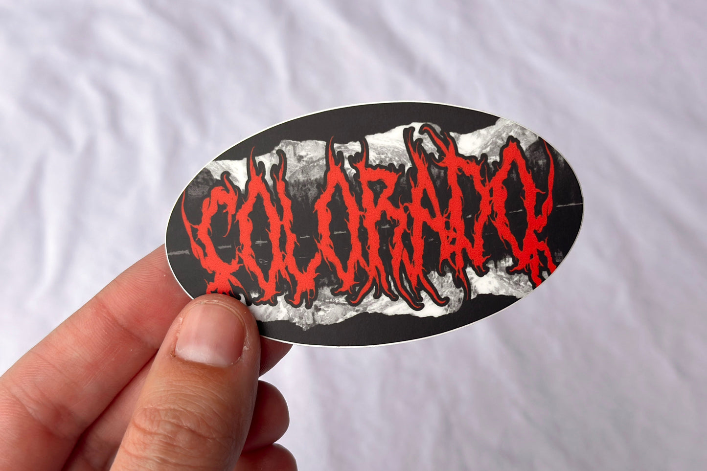 Oval-shaped sticker with red and black 'Colorado' text held by a hand against a white background