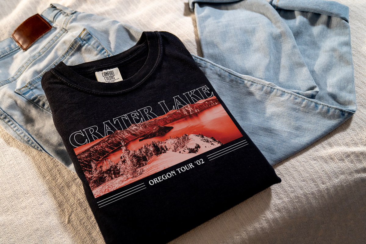 A black t-shirt with a red and white 'Crater Lake' design, laying on jeans