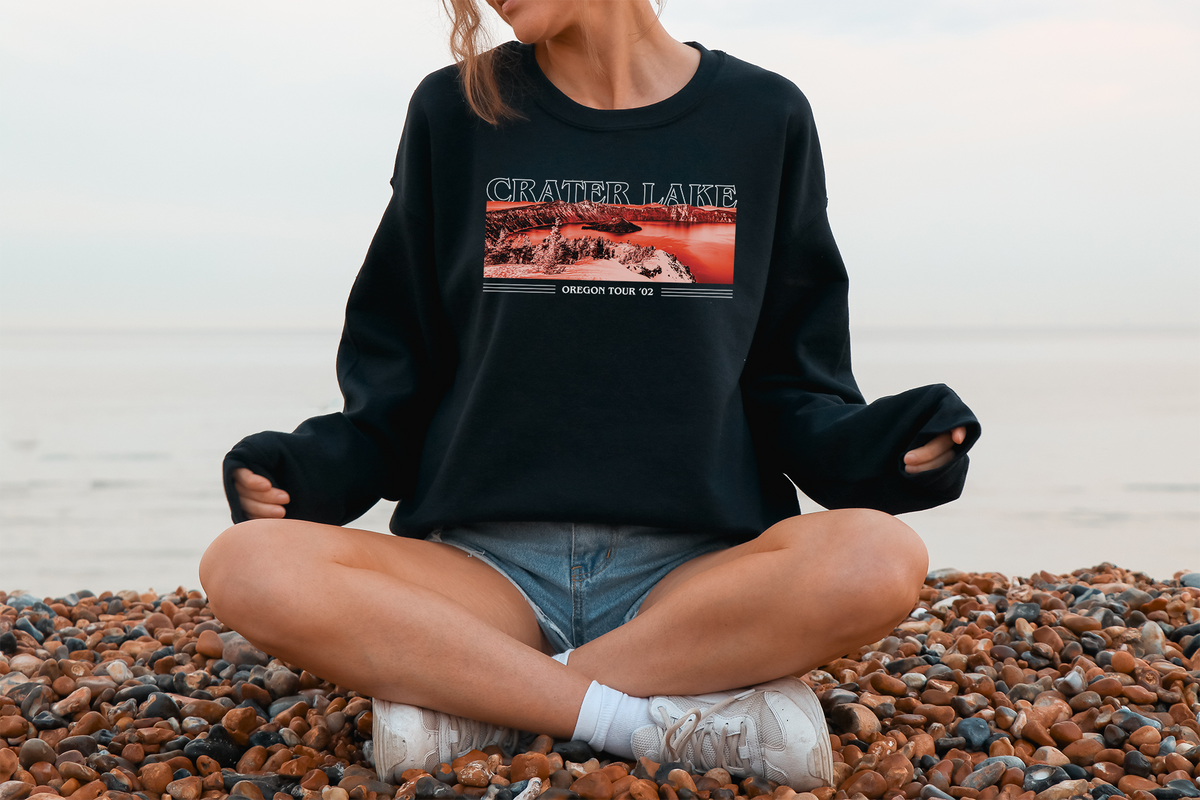 A person sitting on a pebble surface wearing a black sweatshirt with 'CRATER LAKE' text across the chest, paired with blue jean shorts and white sneakers.