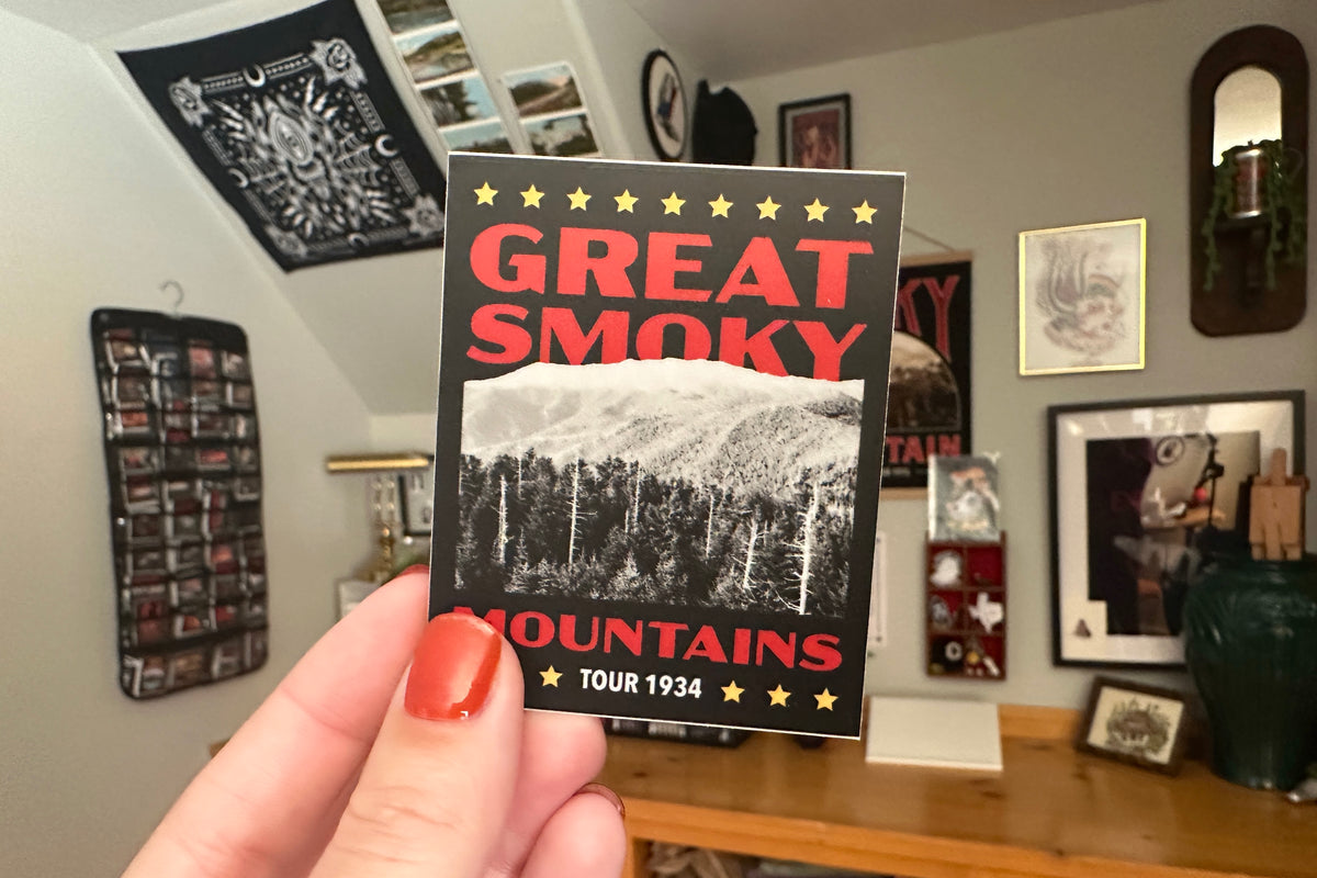 Hand holding a 'Great Smoky Mountains' sticker in a room with framed pictures on the wall.