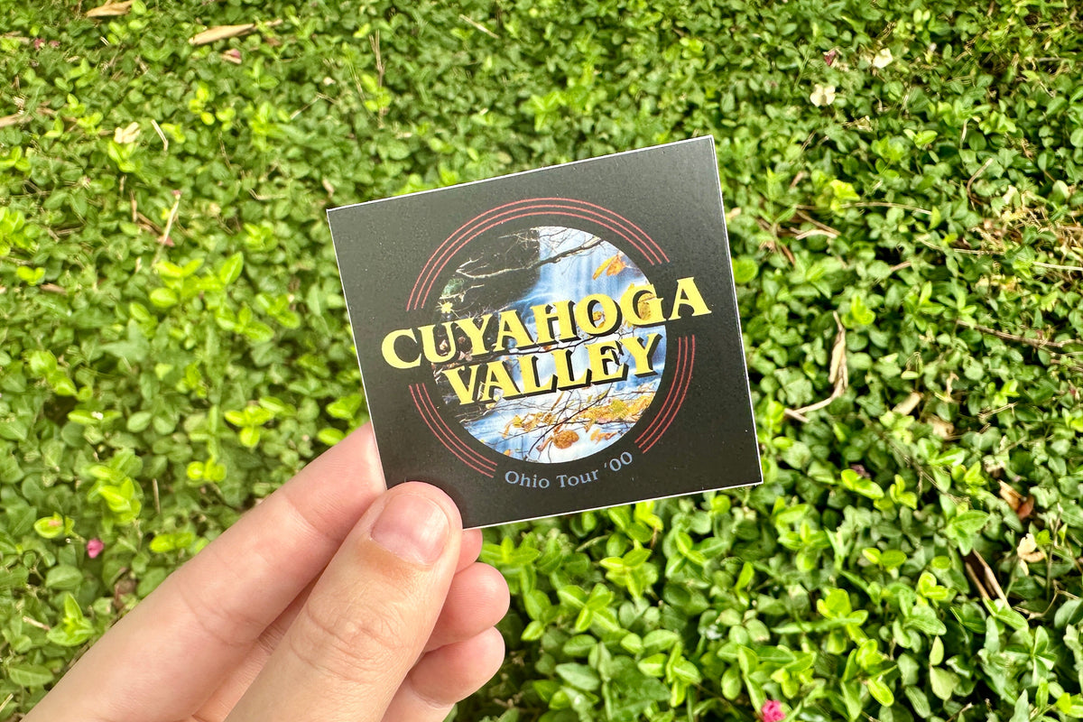 Hand holding a black 'Cuyahoga Valley' sticker against a green grass background