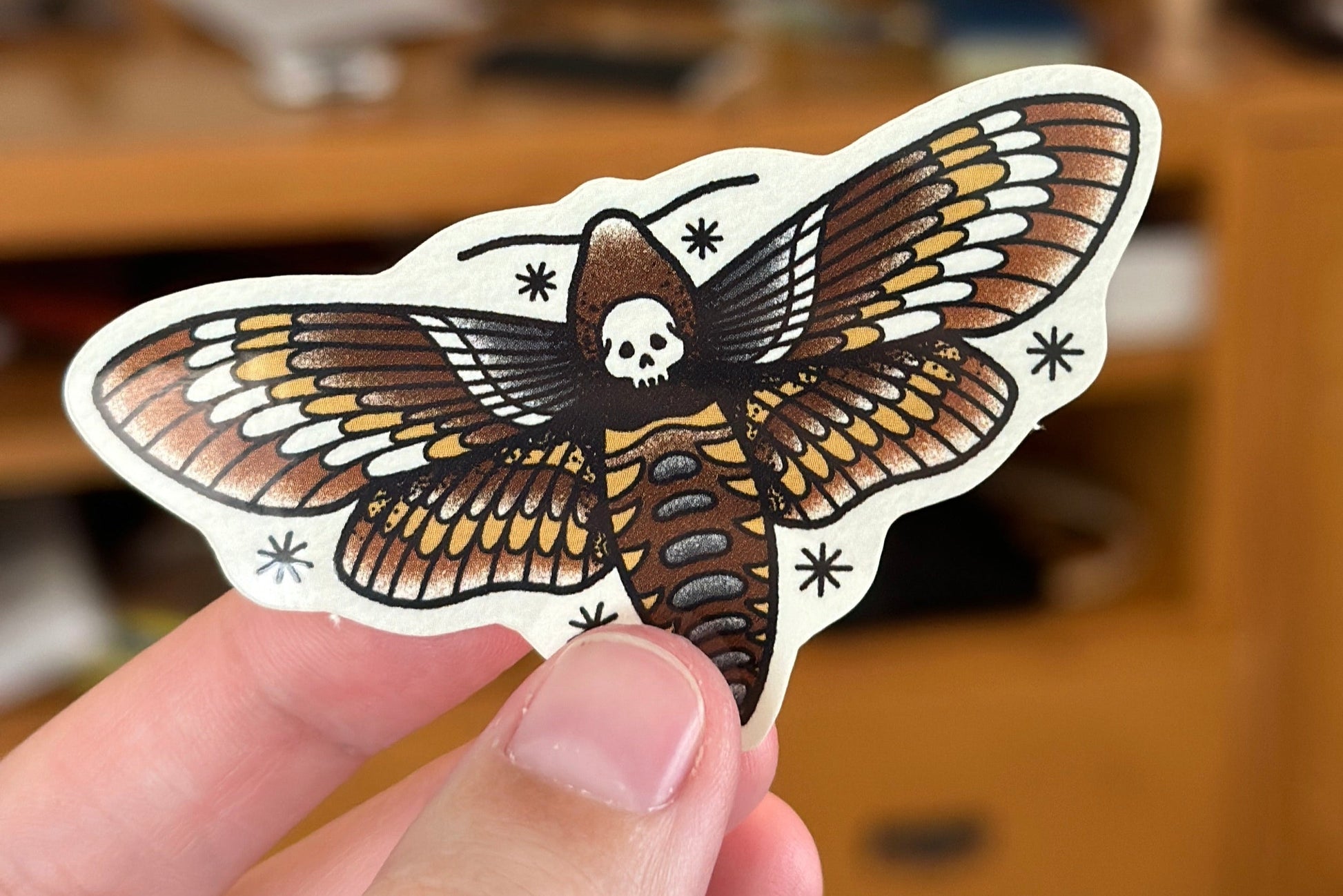 Sticker of a moth with a skull design held by a hand against a blurred background