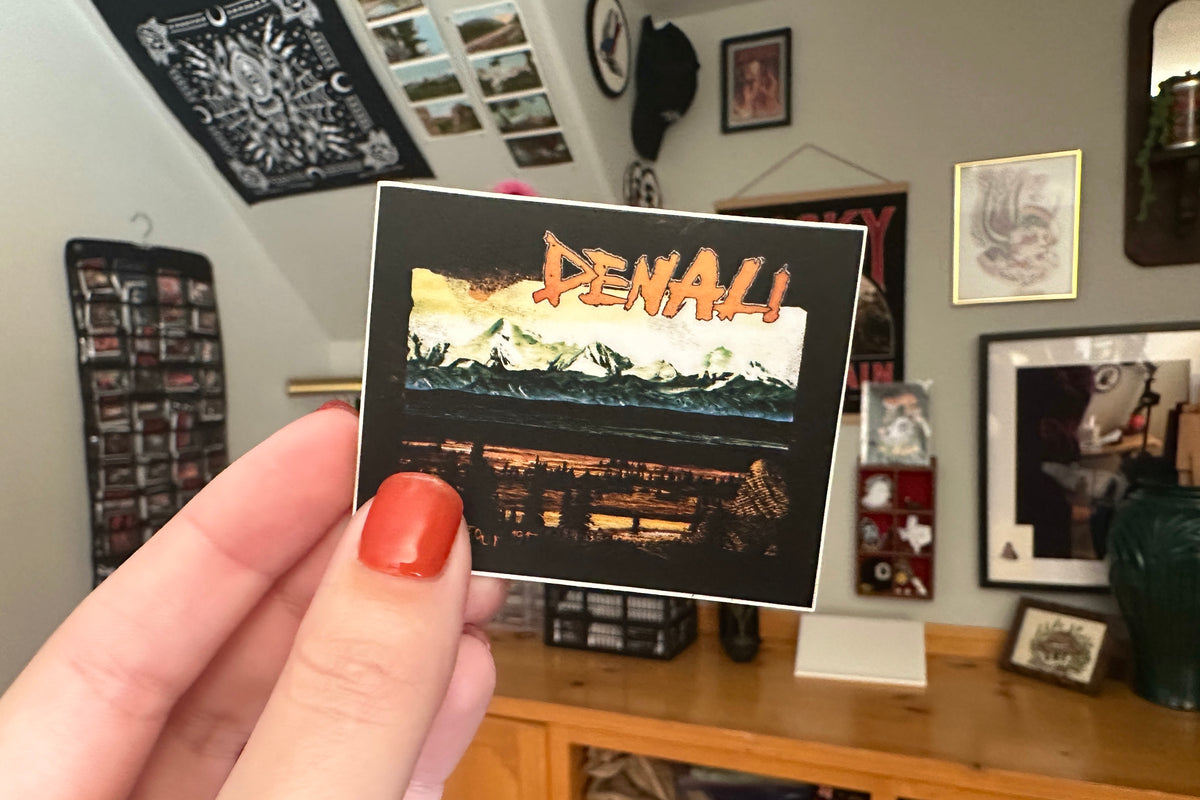 Hand holding a 'Denali' sticker in a room with framed pictures on the wall.