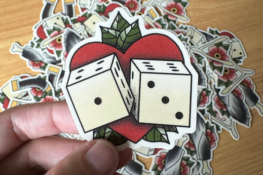 Sticker of two dice on a heart with additional stickers in the background on a wooden surface