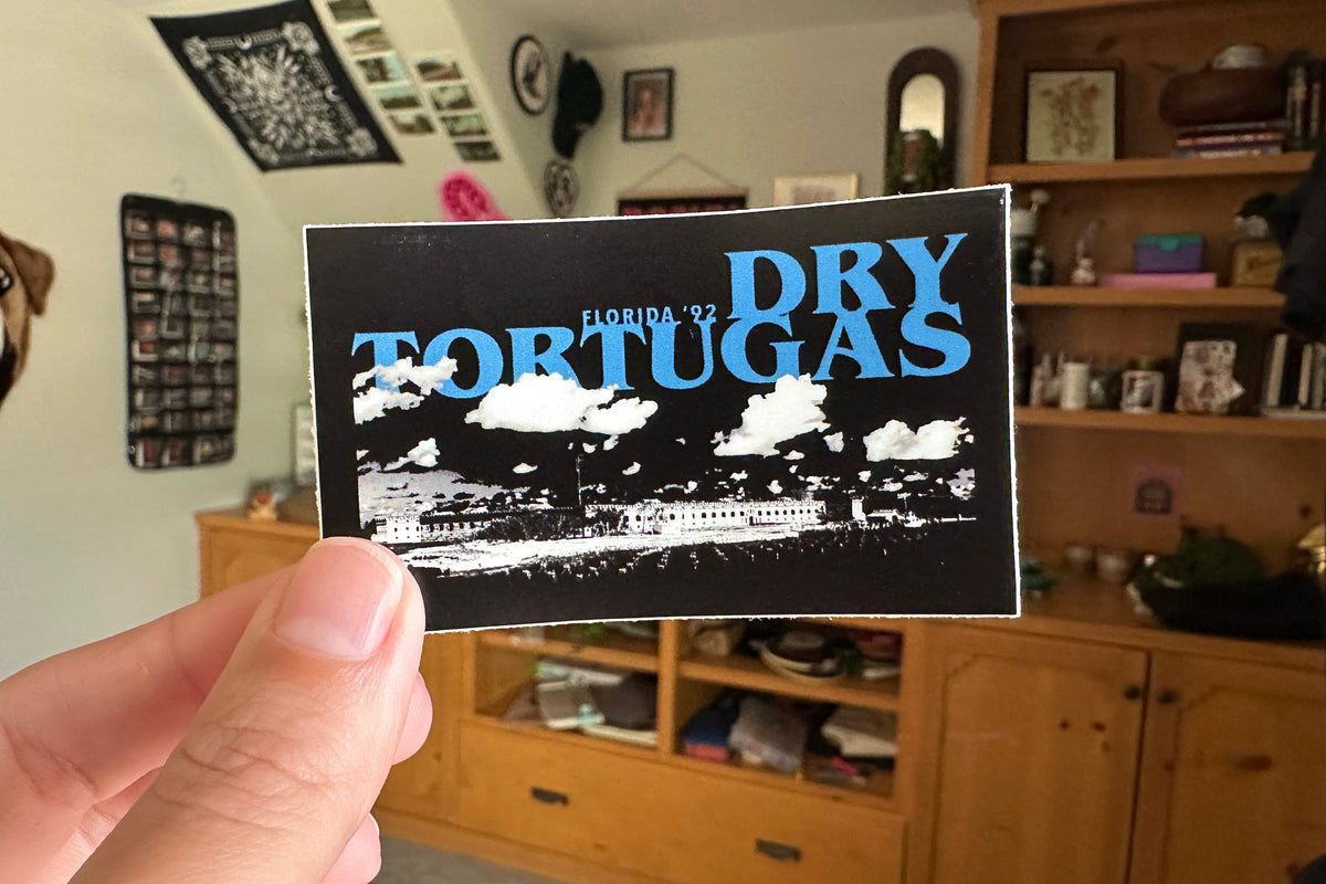Person holding a 'Dry Tortugas' sticker in a room with wooden furniture and decor.