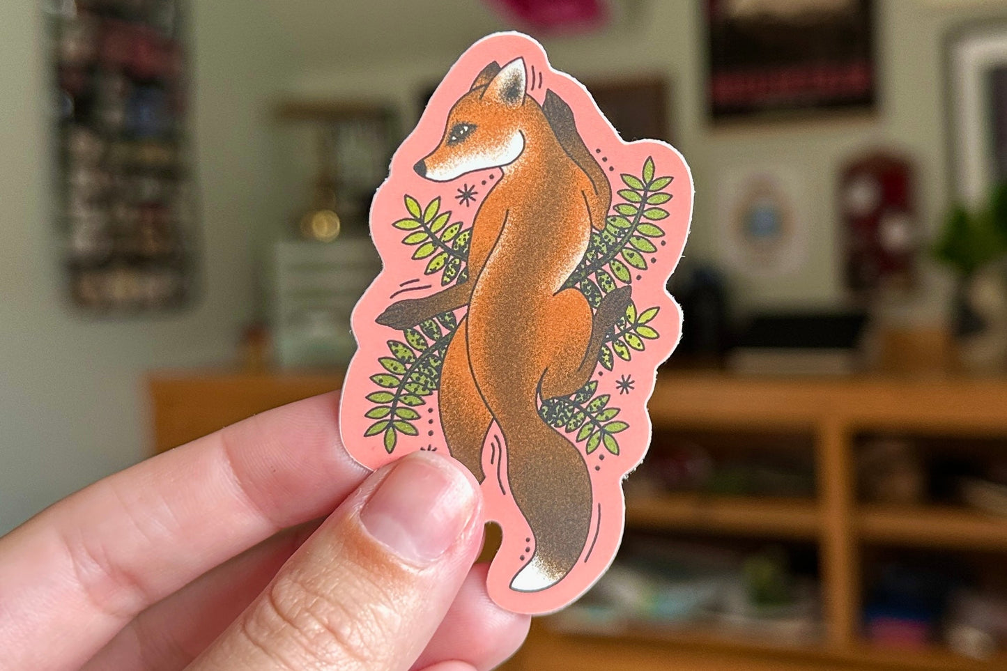 Hand holding a pink sticker with a fox design in a casual indoor setting