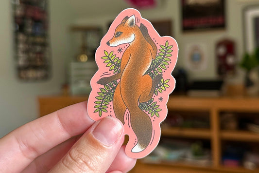Hand holding a pink sticker with a fox design in a casual indoor setting