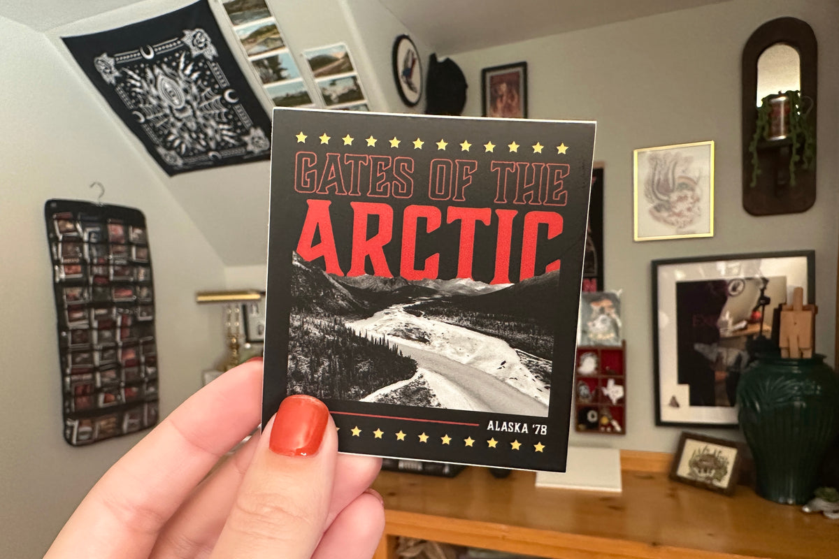 Person holding a 'Gates of the Arctic' sticker with a room in the background.