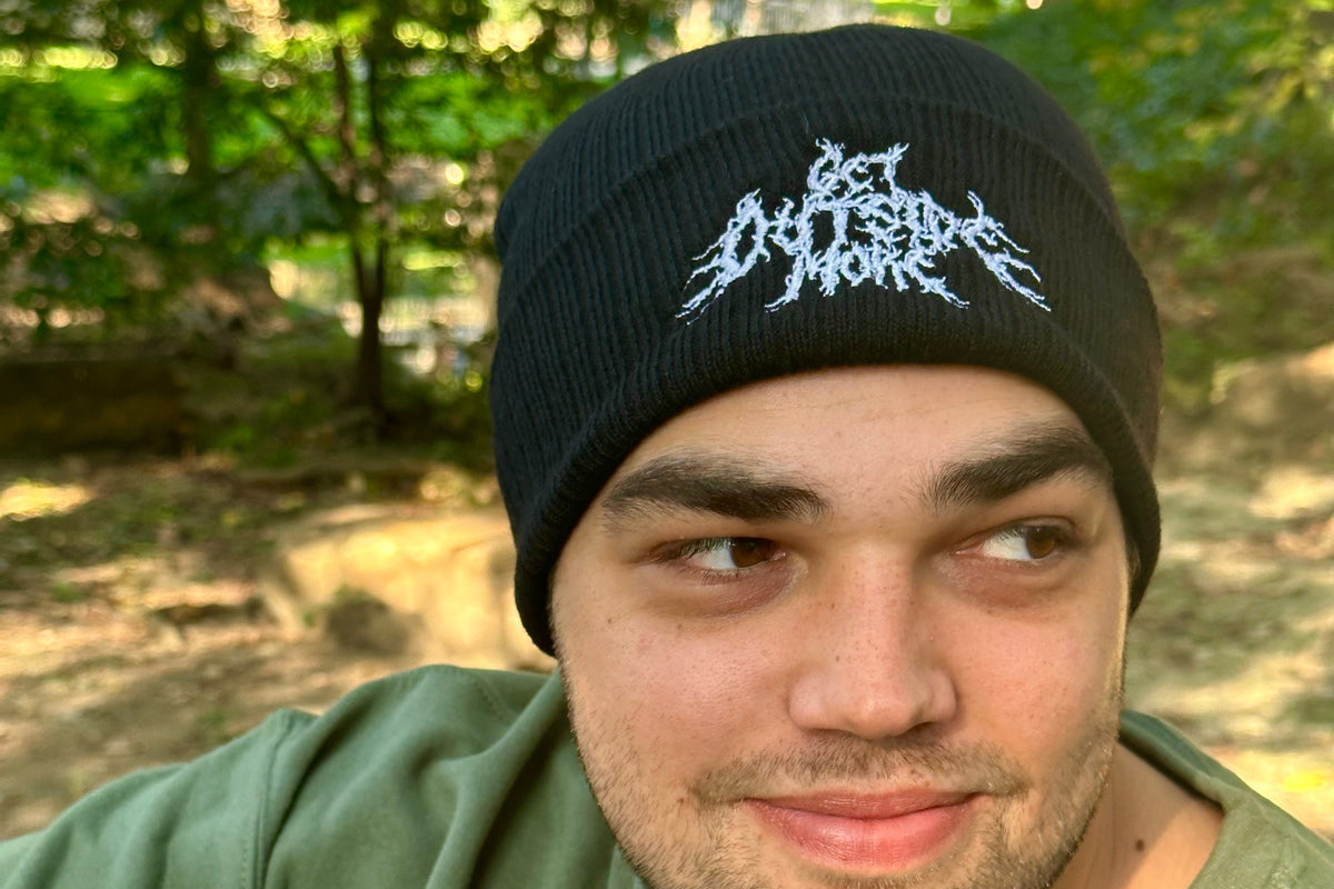 Person wearing a black beanie with white embroidery in a forest setting