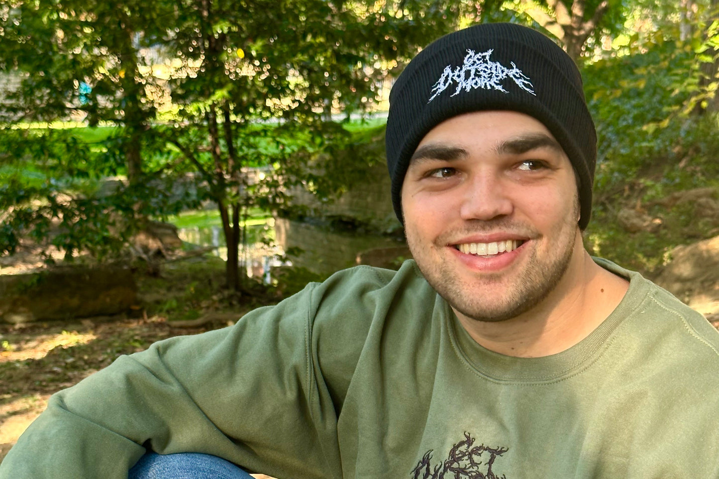 Man wearing a black beanie and green sweatshirt sitting outdoors with trees in the background