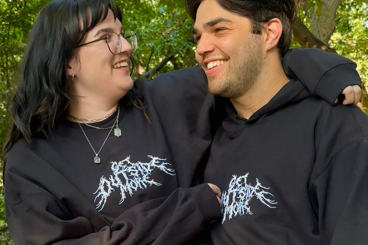 Two people wearing black hoodies with white designs, standing close together outdoors.