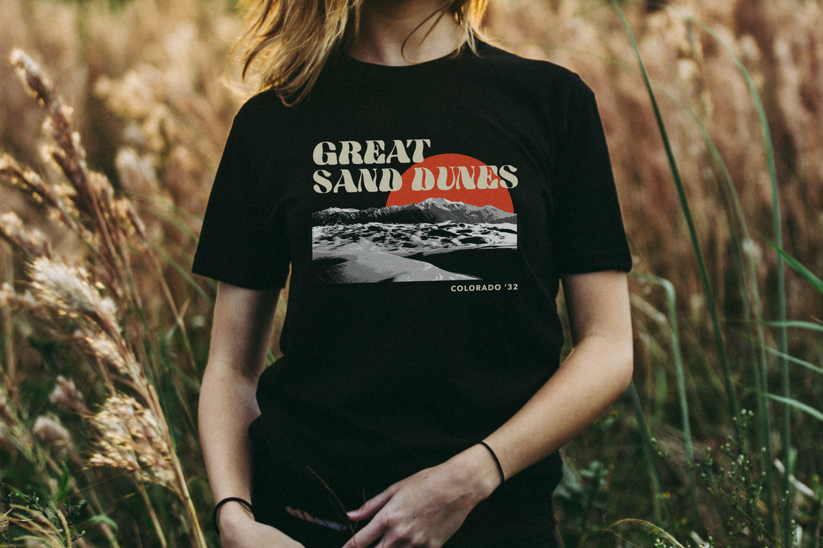 Person wearing a black t-shirt with 'Great Sand Dunes' design in a field