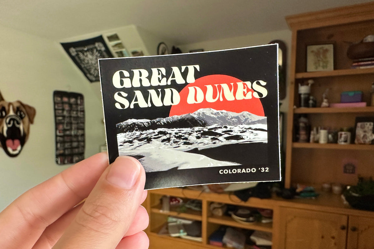 Hand holding a 'Great Sand Dunes Colorado 'sticker in a room with shelves and decor.