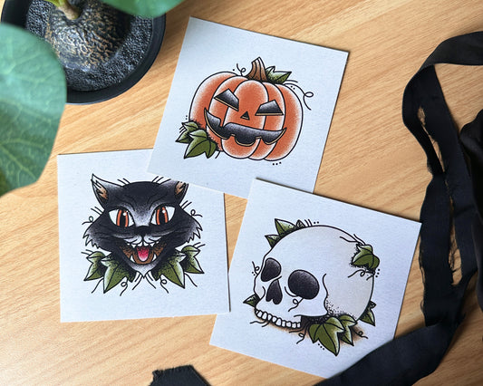 A set of three mini art prints for Halloween, featuring a black cat, a pumpkin with a face, and a skull with a leafy design, laid out on a surface with a black plant and decorative black ribbons.