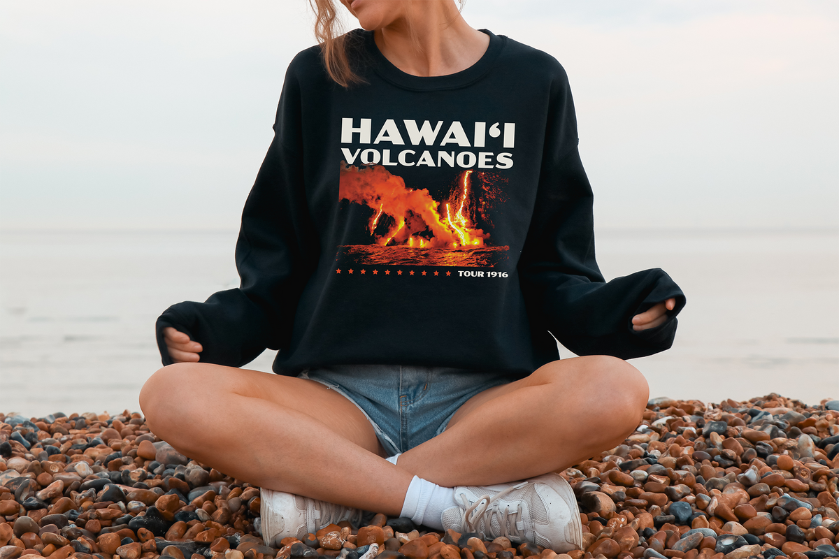 A person sitting cross-legged on a pebble beach wearing a black sweatshirt with 'HAWAII VOLCANOES' printed in white and red text, along with a graphic of a volcanic eruption.