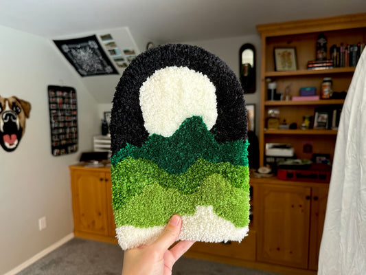 A hand holding a tufted wall hanging rug with green and black colors, featuring a mountain-like design, in a home setting.