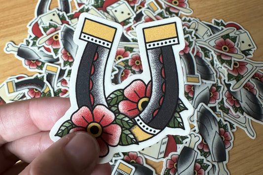 Hand holding a tattoo-style horseshoe sticker with floral designs on a wooden surface
