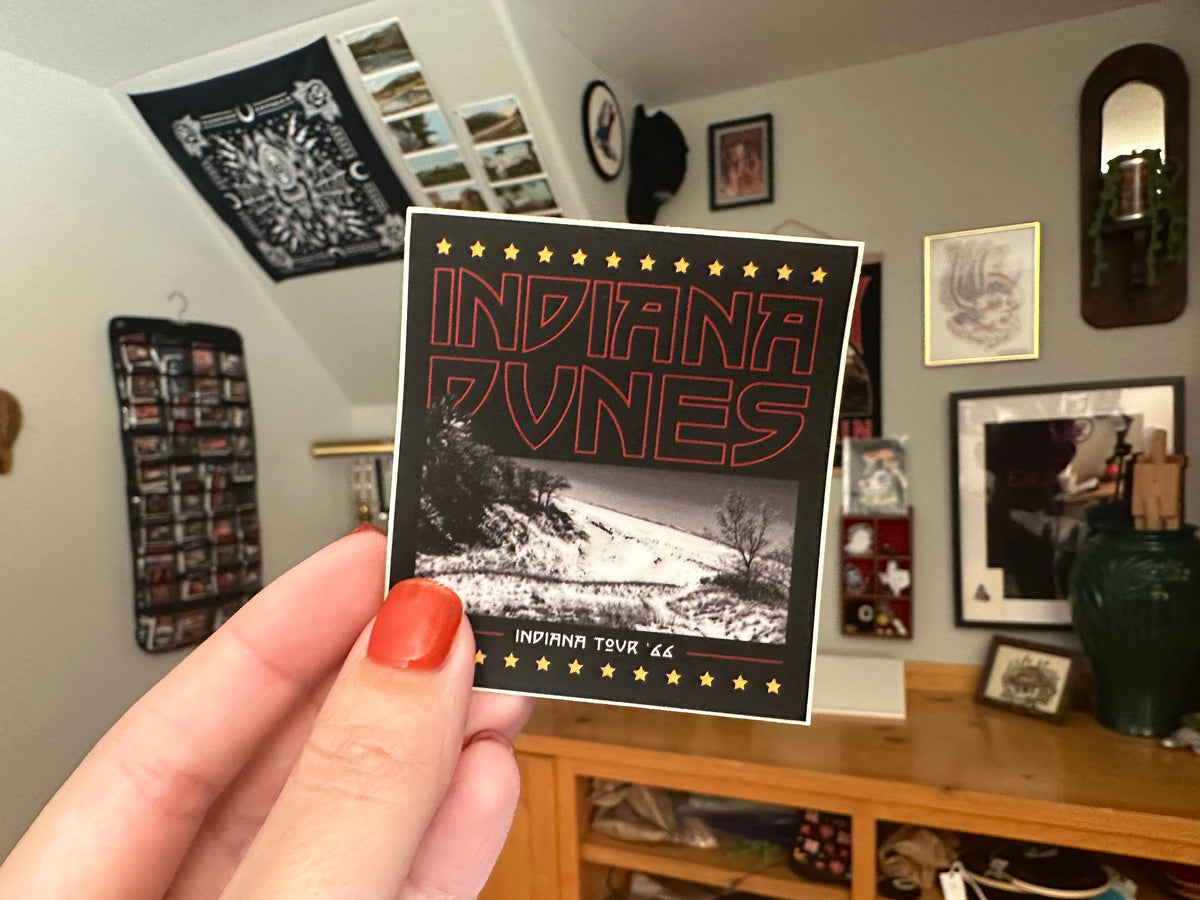 Hand holding a 'Indiana Dunes' sticker in a room with framed pictures on the wall.