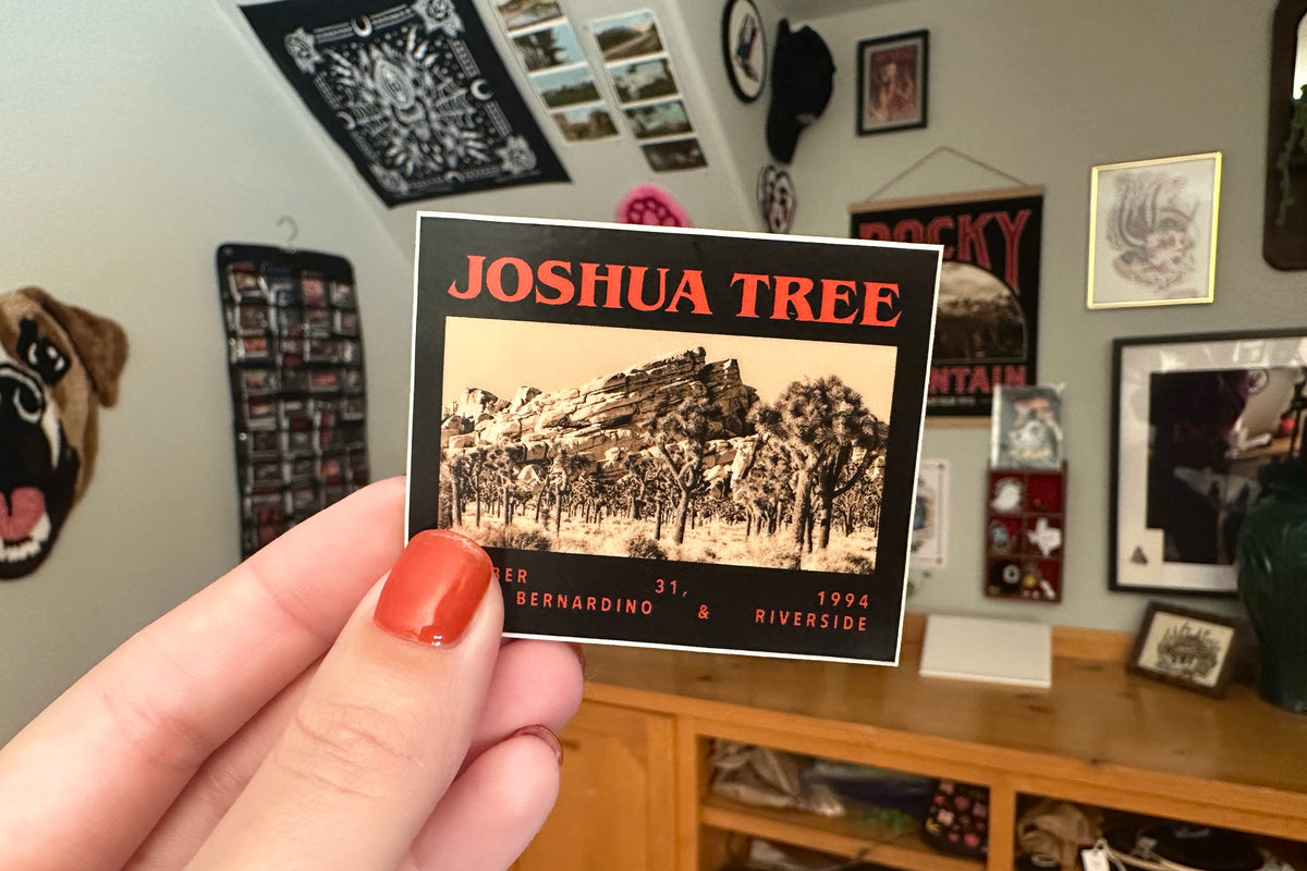 Hand holding a 'Joshua Tree' sticker in a room with framed pictures on the wall.