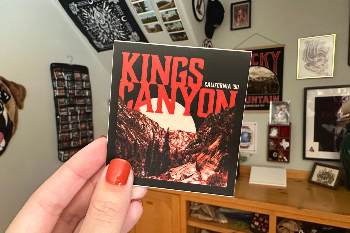 Person holding a 'Kings Canyon' sticker in a room with various posters and photos on the wall.
