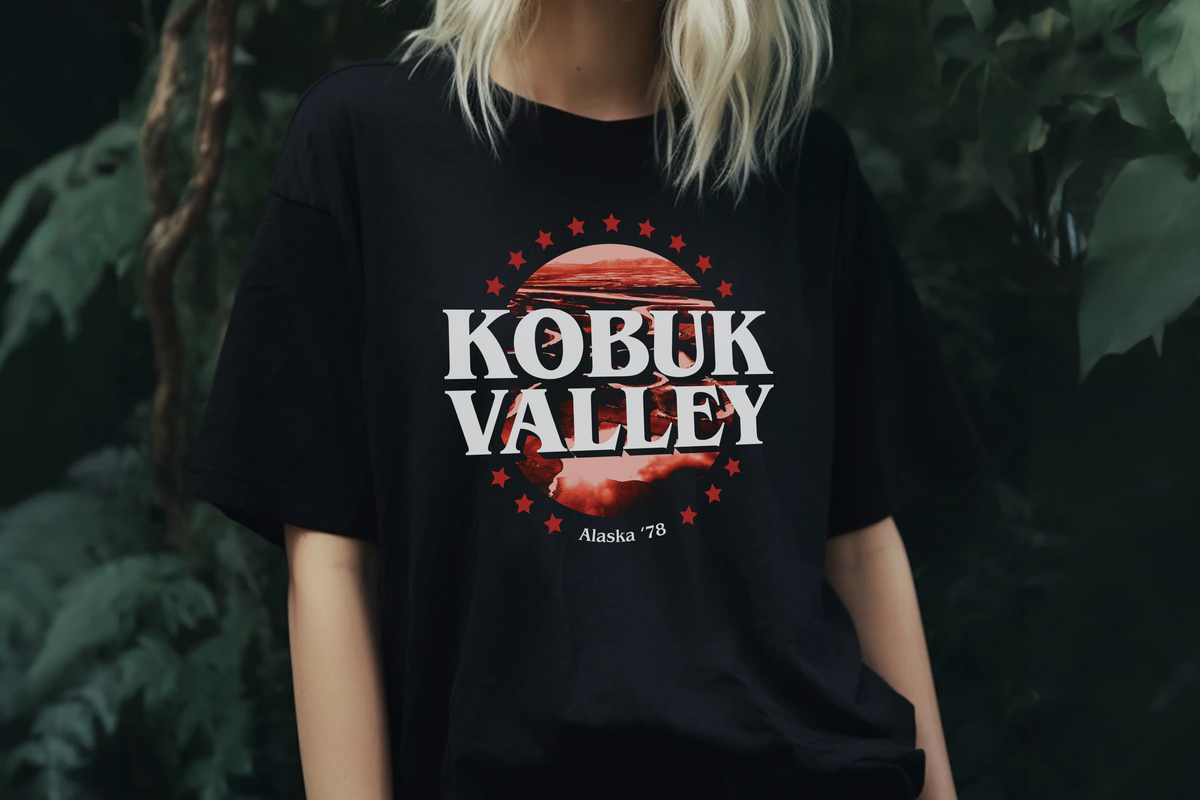 A person wearing an oversized black graphic T-shirt with a red circular design and the words 'KOBUK VALLEY' and 'Alaska -78' printed on the front, photographed against a dark green foliage background.
