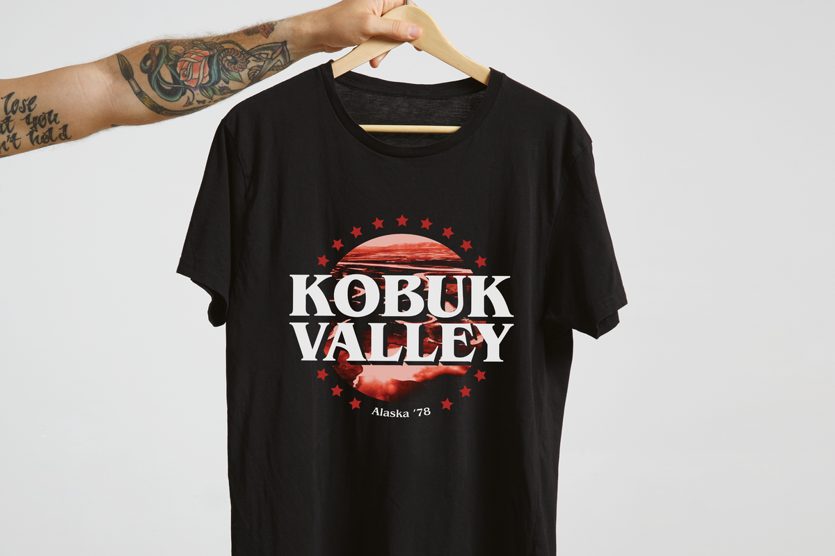 A black short-sleeve graphic T-shirt hanging on a wooden hanger held by a tattooed arm against a light gray studio background. The shirt features a large front graphic with the words “KOBUK VALLEY” in white over a red circular landscape design with small red stars and “Alaska ’78” below.