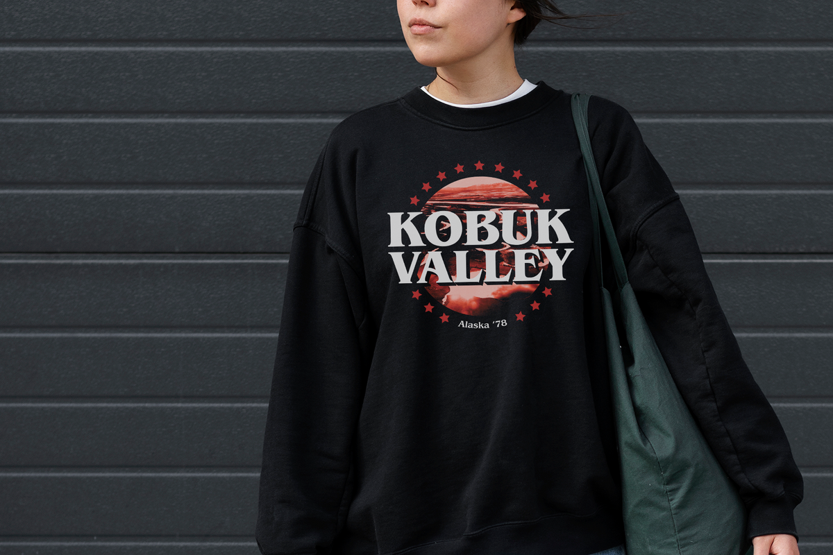 A woman wearing a black crewneck sweatshirt with a 'Kobuk Valley Alaska '78' graphic, paired with a black cap and a green shoulder bag, standing against a dark gray horizontal-slat wall.