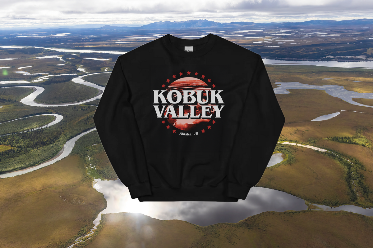 A black crewneck sweatshirt featuring a 'Kobuk Valley' graphic, shown as a digital mockup over an aerial landscape background of winding rivers and marshlands.