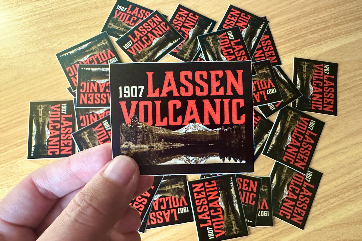 Hand holding a 'Lassen Volcanic' sticker with more stickers scattered on a wooden surface.