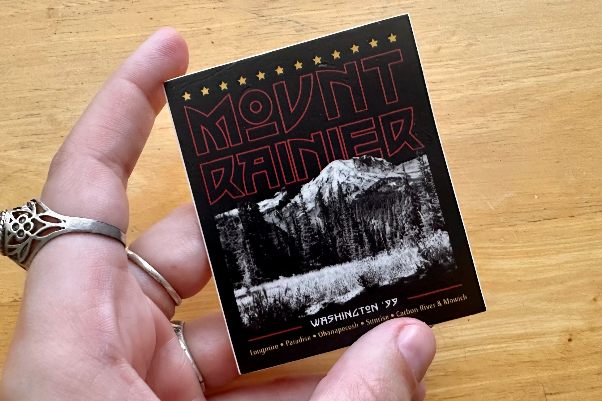 Hand holding a sticker with 'Mount Rainier' text and mountain design on a wooden surface