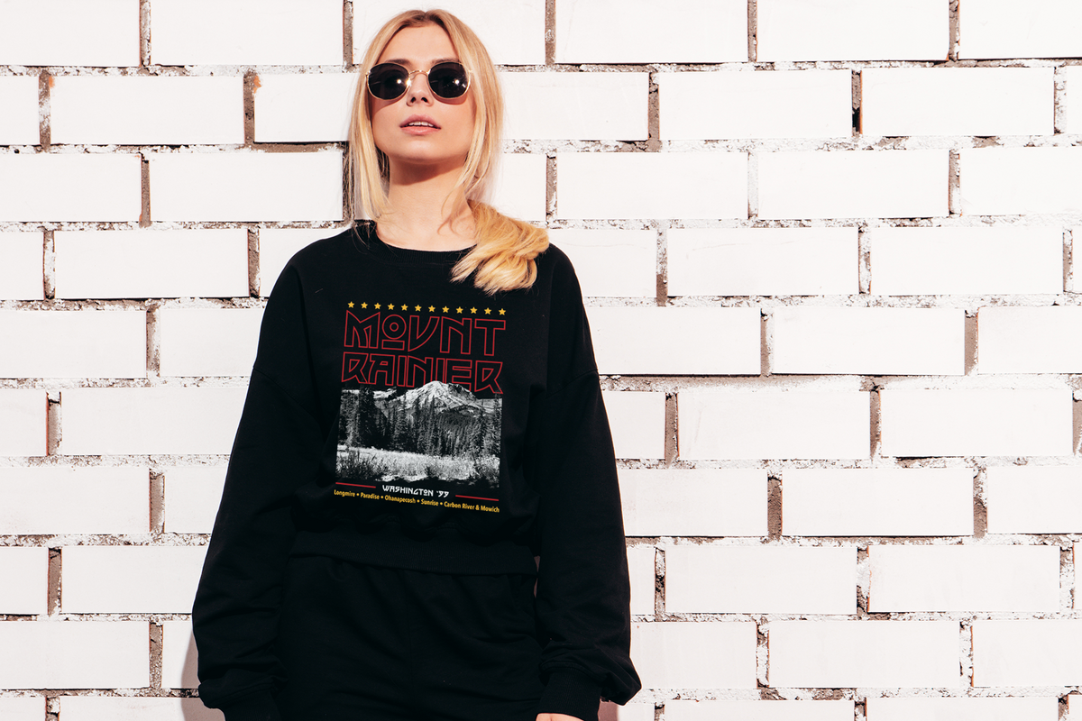 Person wearing a black sweatshirt with a 'Mount Rainier' graphic design against a white brick wall